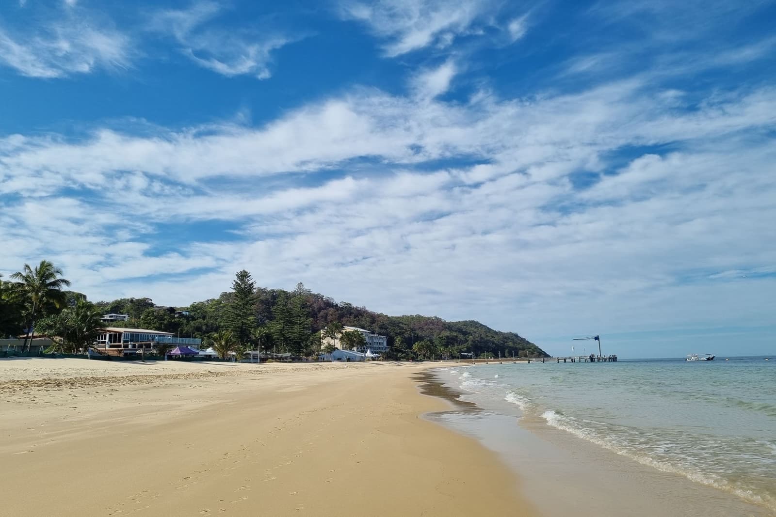 A calm bay beach seen from the sand: pale sand, clear shallows and low buildings along the shore.