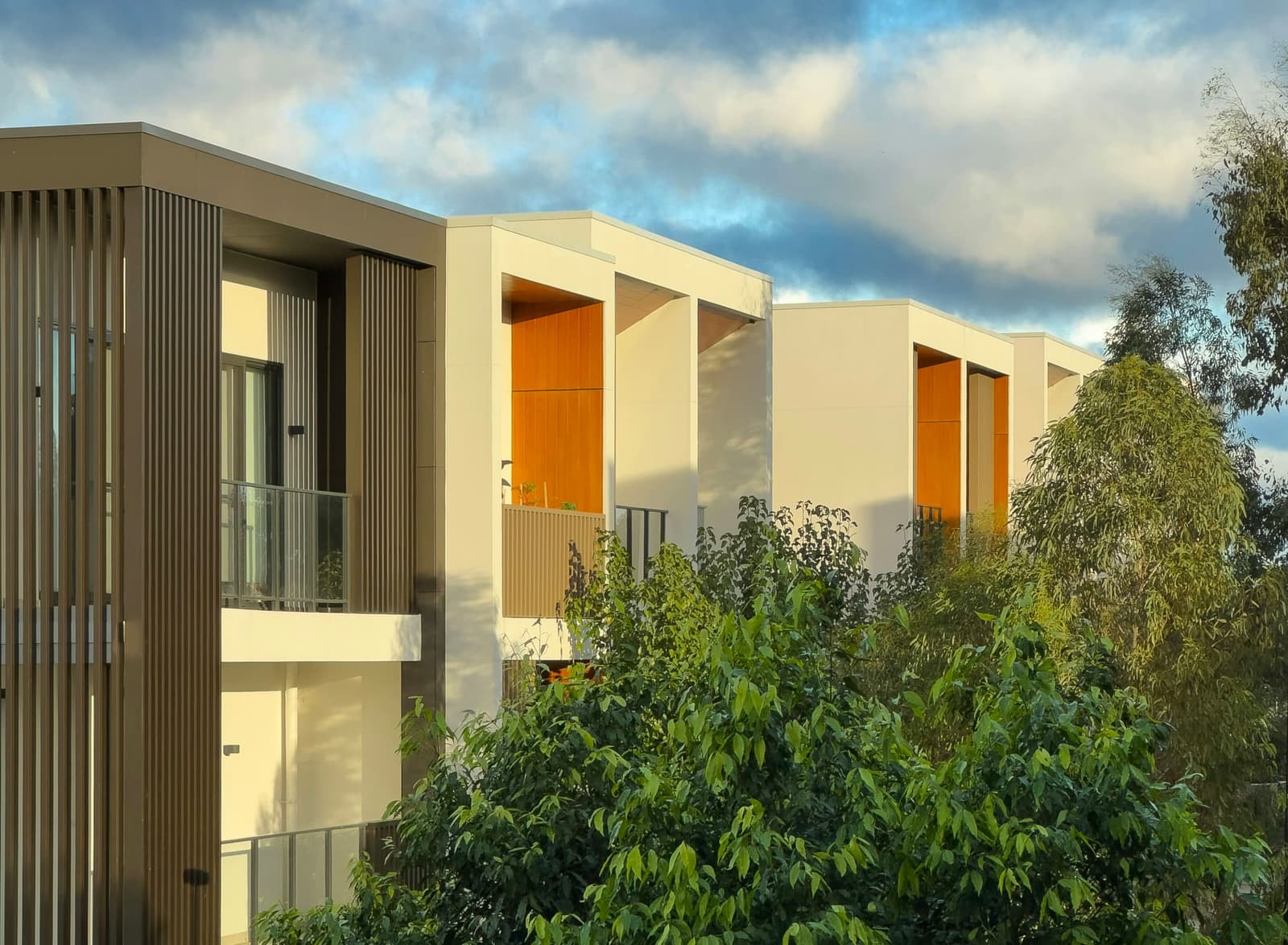 A white-rendered residential building with timber-lined balconies behind gum trees.