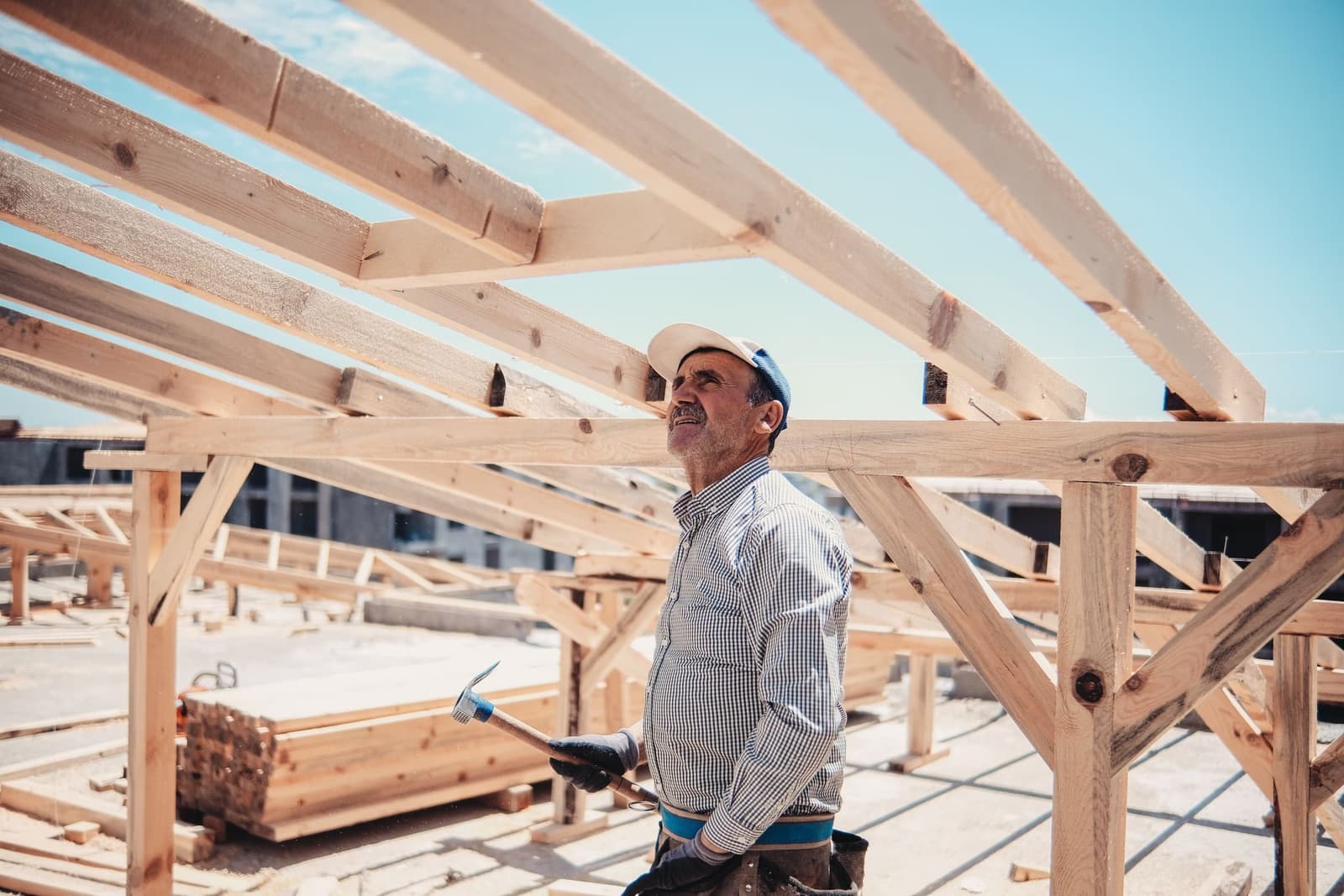 Oliver Harrow in a cap and work shirt, looking up at a timber roof frame against blue sky.