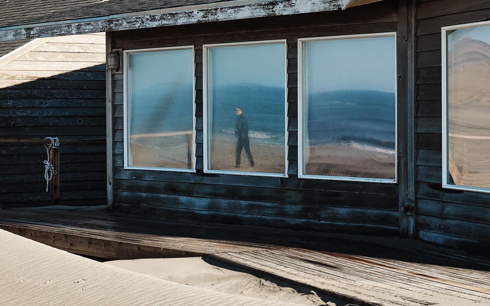 A dark timber house on a dune, its long window reflecting the beach.