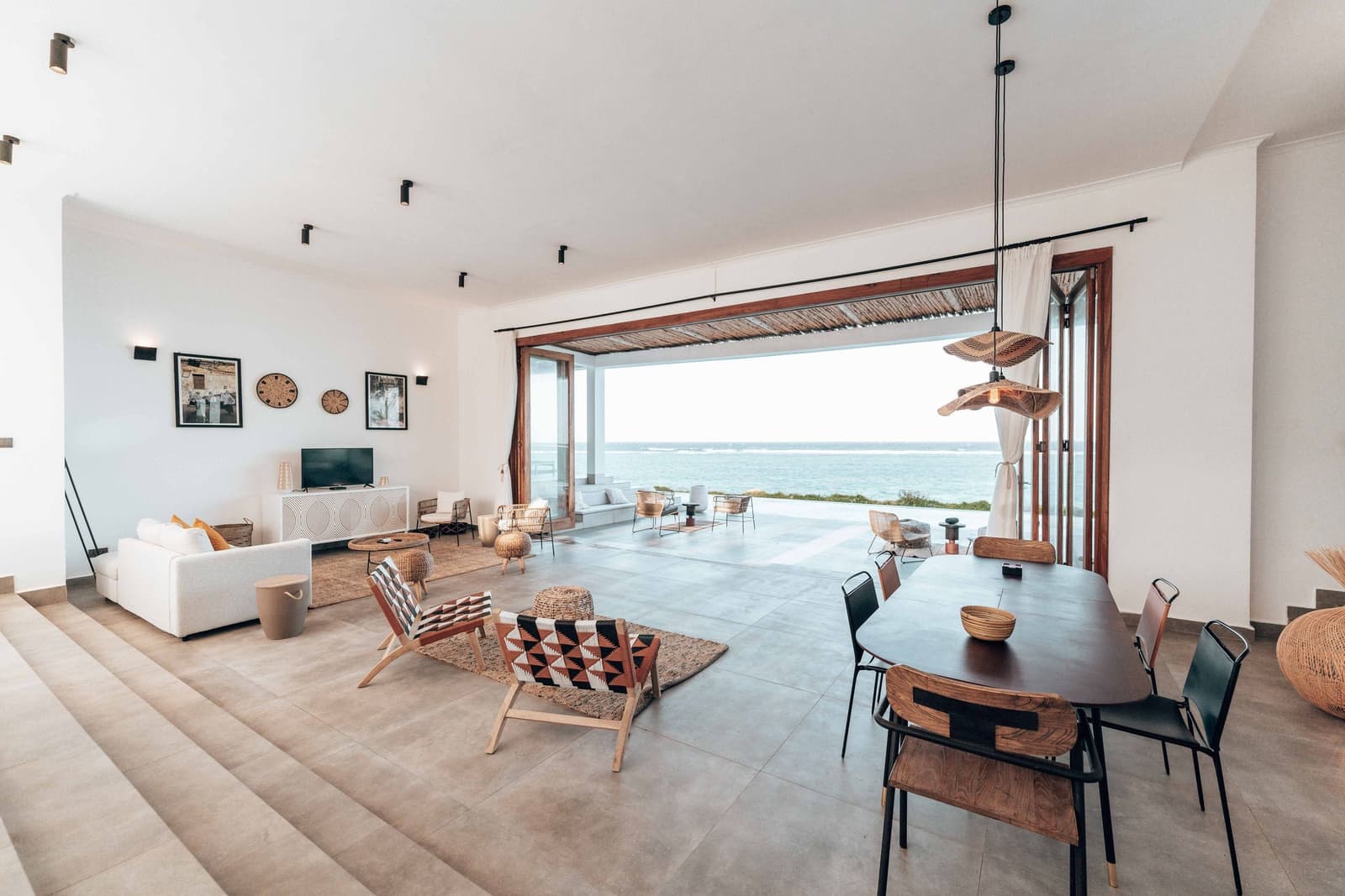 The living room of House 1: white walls, timber and cane furniture, and the bay through a wall of glass.